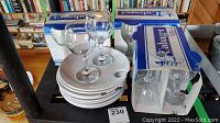 Fourteen 7oz wine glasses including some boxed sets of four labeled Calabajet, with ten white ceramic cheese plates stacked underneath.