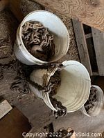 Photo showing two white plastic buckets filled with heavy-duty rusted metal chains, placed on a wooden surface with hay and dirt nearby.