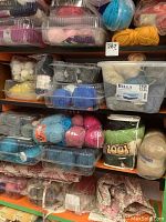 Shelves filled with various partial balls and skeins of knitting wool in plastic containers and bags in different colors and weights.