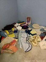 Photo showing multiple scarves, aprons, knit tops, and wigs laid on the floor including one brown wig and various textile items in different colors and prints