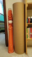 Two rolls of packing paper standing vertically next to each other; one large brown kraft paper roll and one smaller white roll wrapped in orange protective covering near a bookshelf.