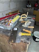 Various painting and drywall tools arranged on a wooden surface including paint brushes, roller, metal paint tray, and several hand tools.