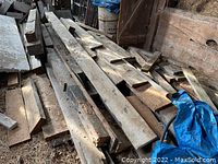 Pile of rough cut hardwood boards stacked inside barn, various lengths and widths, showing rough unfinished surfaces with dirt and debris.