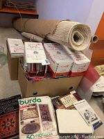 Overview of rolled jute/linen backing and boxes of rug hooking wool with pattern books visible on table.