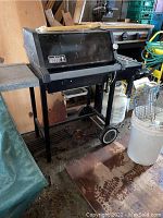 Full view of the Weber BBQ grill showing metal frame, side trays, wheels, and propane tank underneath. Some items stored on top.