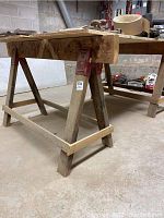 Side angle of one wooden saw horse showing wood construction, red metal brackets, and the overall shape and structure including legs and cross brace.