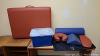 Photo of folded brown vinyl foam massage bed with handle, plastic container with white lid, black transport bag and various cushions including blue and brown vinyl