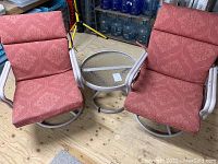 Image showing two swivel patio chairs with patterned red cushions and a round glass top coffee table on a wooden floor.