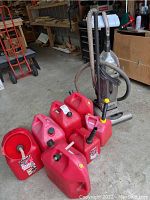 Photo showing seven red plastic gas containers with different sized spouts, positioned on floor along with an upright Hoover Wind Tunnel vacuum cleaner.