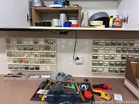 Overall view of workbench with Craftsman vise, assorted hand tools including hammers, measuring tapes, utility knife, and small parts storage boxes attached to the wall filled with screws and nails.