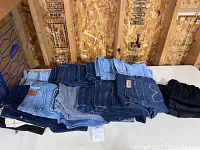 Overview of the folded jeans pile showing a mix of denim colors and black bottoms on a table against a plywood wall.