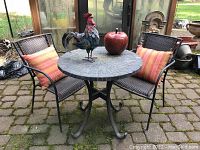 Full view of bistro table with two chairs having orange striped cushions, decorative metal rooster and large glazed apple on table