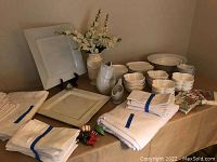 Table display showing various white and cream tablecloths, napkins, and dishes including platters, bowls, gravy boat, and decorative vase with floral arrangement.