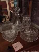 Wide view showing heavy leaded cut crystal bowl, decanter with stopper, pressed glass butter dish, and vase, all arranged on wooden surface inside cabinet.