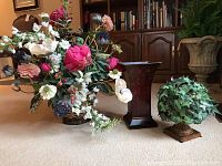 Full lot showing the large silk flower arrangement in rattan basket, metal tole planter/urn, and moss topiary sphere with urn base, set against a living room backdrop.