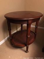 Front view of oval end table showcasing wooden top and rattan lower shelf, tapered legs and wheels visible.