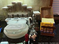Wide view of set of various food storage containers, glass and plastic, along with salt and pepper shakers, decorative items, napkin holder, and maple syrup pitcher on kitchen counter