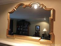 Front view of large gold gilded scalloped wood framed mirror reflecting a room with bookshelf, lamp, and chairs.