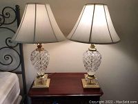 Pair of cut glass and brass table lamps with white silk shades sitting on wooden furniture beside a metal headboard.
