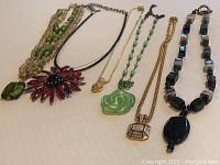 Full lot showing five necklaces including the Swarovski multi-strand green bead necklace, red floral pendant necklace, green rose medallion pendant necklace, geometric jeweled pendant necklace, and black and gray stone beaded necklace.