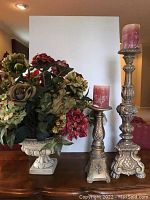 Full view of plaster urn with floral arrangement and two gilded plaster candlestick holders with candles on wooden surface against white backdrop.