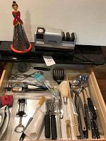 Photo showing Chef's Choice 100 electric sharpener, triangle-shaped grater with woman figure on top, and opened drawer containing various kitchen utensils and organizers.