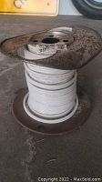 Photo of partially used metal spool with white insulated electrical wire wound around it, label showing wire gauge and length.