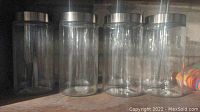 Front view of four empty clear glass cylindrical containers with silver metal lids arranged side by side on a shelf.