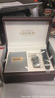 Open presentation box showing Gruen wristwatch with metal band and additional black leather strap inside, accompanied by instruction booklet.