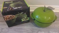 Green cast iron enamel apple pot beside its original box showing the Lodge branding and apple pot image.