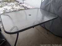 Rectangular glass top outdoor patio table with black metal frame and legs, photographed on a concrete patio with snowy backyard visible.