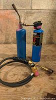 Two propane torches with blue propane cylinders and brass nozzles standing upright on wooden surface.