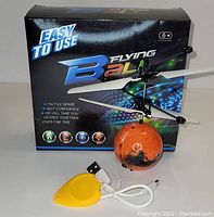 Red spherical flying ball helicopter toy with black dual rotor blades, yellow remote control, and white USB charging cable in front of the product box.
