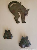 Photo of metal cat sculpture with arched back and two separate cat face plaques placed below it on a light surface.
