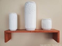 Wooden floating shelf holding three white ceramic vases with embossed floral design: tall cylindrical, medium cylindrical, and short square shape.