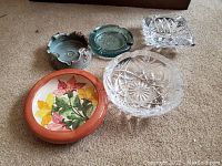 Photo showing five assorted ashtrays on carpeted floor: clear cut glass with star pattern, clear textured cut glass with rectangular pattern, green textured ceramic, greenish glazed ceramic with scalloped edges, and round ceramic with floral design.