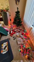 Overall photo showing a 4-inch artificial Christmas tree, multiple strands of multicolored Christmas lights, assorted ornaments spread on the floor, and a large green storage box.