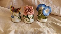 Set of three decorated porcelain bells displayed on a cloth background showing all three designs side by side: butterfly and blue flowers, pink rose, blue morning glory.