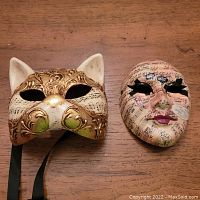 Two masks on wood surface: cat-shaped gold and white mask with music notes, and full-face mask covered in printed musical notes with lashes and lips.