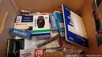 Top-down photo of a cardboard box containing assorted office supplies including paper, envelopes, stapler, staples, hole puncher and notepads.