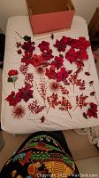 Multiple red botanical Christmas ornaments including poinsettia flowers, glittery leaves, and spiky floral decorations laid out on a white bedspread with a box in background.
