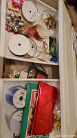 View of white storage box drawer containing multiple spools of ribbon, assorted bows in gold and metallic colors, and some loose ribbons and bows.
