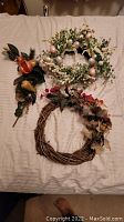 Photo showing two seasonal wreaths and one decorative tree bough with artificial fruit laid out on a bed.