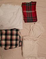 Photo of folded table cloths and napkins showing varied plaid and solid patterns, with white jacquard, cream, red and green plaid fabrics.