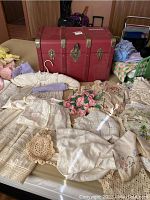 Red faux-leather American Girl doll trunk with metal hardware and several vintage lace and embroidered textiles and doll dresses arranged around it on a table.