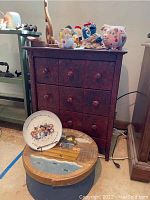 Photo of a dark wood cabinet with multiple small drawer fronts, assorted decorative figurines on top including a piggy bank, and a round wooden decorative box in front.