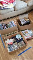 Four cardboard boxes on a hardwood floor containing assorted CDs, DVDs, video games, and game controllers.