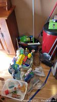 Overview of cleaning supplies including trash can, brooms, mop bucket, and various cleaning fluids on floor next to cabinet.