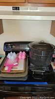 View of stove with large black speckled pot, stacked metal baking sheets, pink cutting board, and Jell-O silicone molds on top.