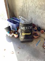 Wide shot showing a variety of planters in different sizes, the opened large bag of black mulch, and multiple small pots grouped together near a wall in natural light.